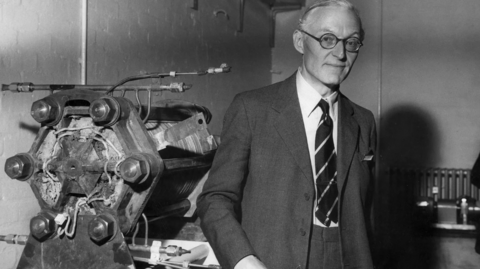 Tom Bacon is seen in an old black and white photograph standing next to the fuel cell he developed. He has receding fair hair, dark-rimmed round spectacles and is wearing a a dark suit, a white shirt and a striped tie