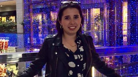 Marta Elena Vento poses for a photograph on a balcony in a retail centre. She wears a black jacket and a patterned black and white dress.