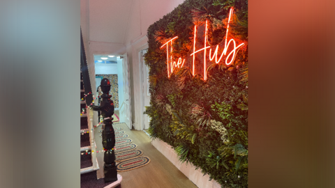 Red illuminated lettering which says 'The Hub' sits on a wall feature of greenery inside a hallway 