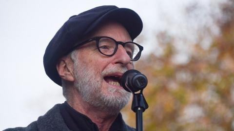 Billy Bragg, wearing a black flat cap and glasses, sings into a microphone. He is also wearing a grey woollen coat with a red button. He is outside and there is a tree behind him.