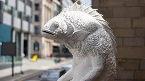 A close-up of the sculpture Skalli which is white in colour and located on a plinth at the side of Liverpool Parish Church. It is in an elevated position with a road with traffic passing buy below.