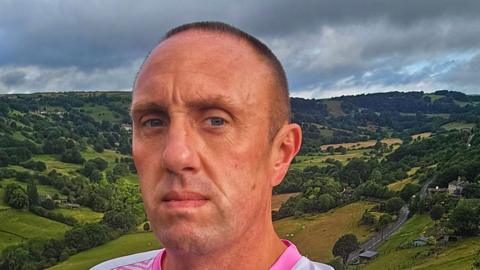 A man with very short dark hair, wearing a sports top, stands in front of a countryside landscape.