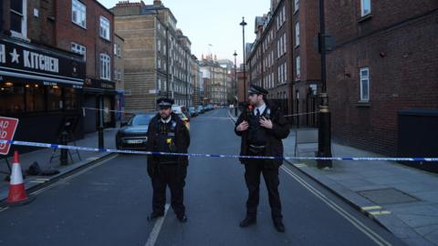Two police officers stand behind a police cordon on a street in London