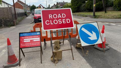 a red road closed sign with other signs and traffic cones in the middle of a road with a strip of fresh tarmac