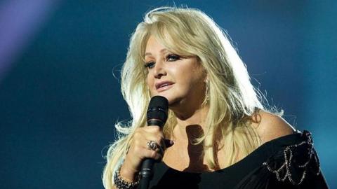 Bonnie Tyler, wearing a black dress, can be seen singing on stage