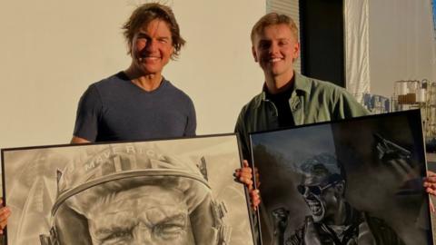 Actor Tom Cruise standing next to artist Wilf Elliott. They are outside and standing in front of a white wall, both holding framed charcoal portraits of Cruise.
