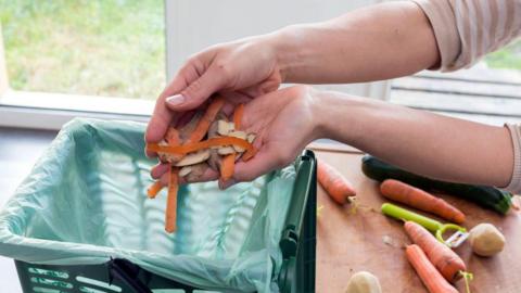 A person holding small potato and carrot peelings above a small green food bin, with a green liner in it. On a chopping board next to the bin are carrots, potatoes and an courgette