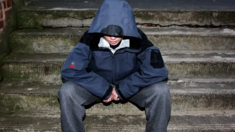 A young boy sits on some steps with a hood up, looking at the floor. He has jeans on and his face is partially hidden.