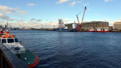 A view over the River Tees of Port Clarence. A number of vessels are docked at the port and a crane towers above them.