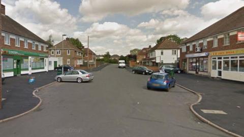 Street view of Sun Way in the Braunstone area of Leicester showing shops and cars to the left and right of the road.