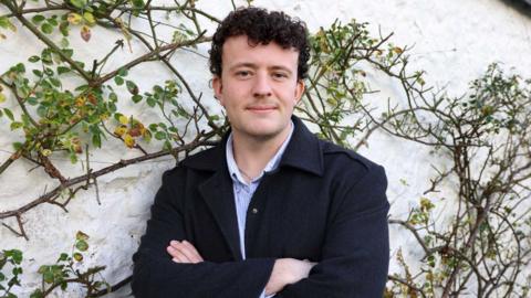An image of a man in a black suit jacket in front of some greenery