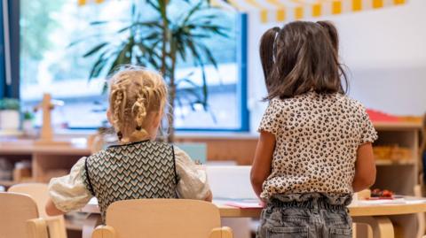 Two nursery children sit and stand at a table playing.