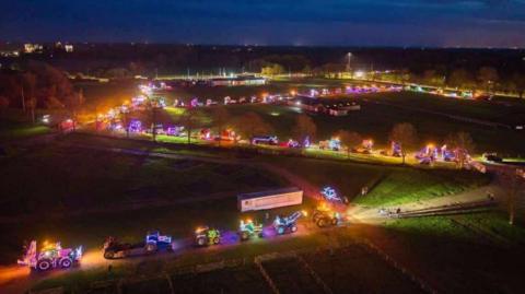 A drone shot of brightly-lit trucks and tractors snaking along a road that zig-zags through green fields, with the lights stretching into the middle distance under a dark-blue evening sky.