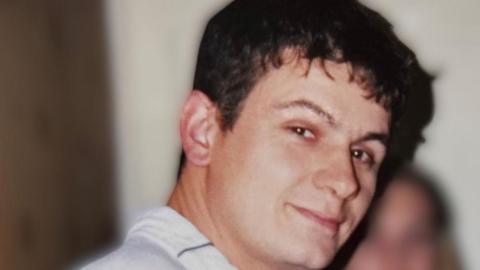 A man with short dark hair turns to the camera. He is wearing a white shirt.  