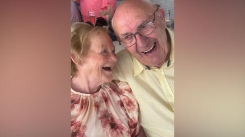 An elderly couple embrace. She has light hair and a floral top. He has grey short hair and glasses. He wears a yellow shirt. They are both smiling