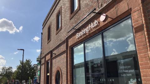 A red brick bank building on a high street with a sign that says 'Banking hub'. The sky is blue in the distance with a few clouds. 