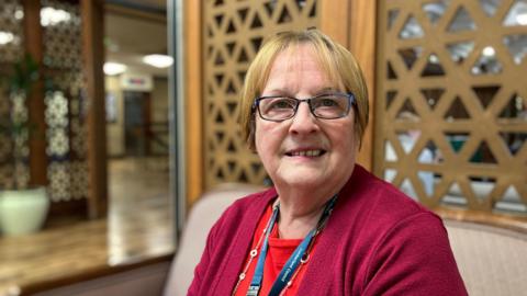 Cumberland Council finance portfolio holder Barbara Cannon. She has short blonde hair and blue eyes. She wears glasses with a blue rectangular frame. She is wearing a burgundy cardigan over a red top. She is wearing a necklace and a lanyard around her neck.
