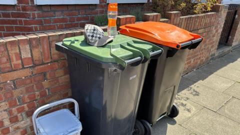 Two large outdoor wheelie bins placed on a pavement beside a low brick wall in front of a house. One bin has a green lid and the other has an orange lid.