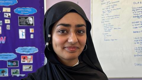 Areebah smiles at the camera as she stand is front of a white board covered in writing. Her head is covered with a black hijab. 