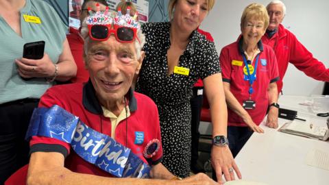 A man wearing a red top with a birthday banner has birthday glasses on his head, as colleagues look on behind him. 