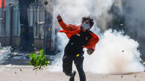 An Indonesian protester wearing a red jacket, black trousers, a face mask and clear goggles hurls a rock as a tear gas canister explodes in a cloud of white smoke behind me