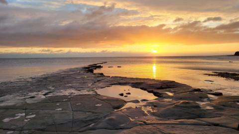 The sun is setting over the sea casting a golden glow over the sky and water beneath it. In the foreground are rocks and rock pools created by the tide.