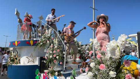 A view of one of the flower displays at the Battle of Flowers parade. There are men playing guitars and a woman in a p;ink dress wearing a cowboy hat is singing into a microphone. There are sprays of pink and white carnations and white gypsophila all over the float.