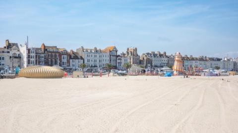 White sandy beach with attractions along its edge and buildings stretching behind.  It is a bright, sunny day.