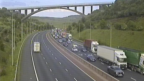 Grainy traffic camera image showing a build-up of traffic in the eastbound lane, while quieter heading westbound. There is a bridge over the motorway.