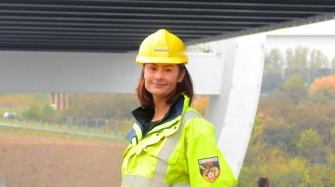 Amy Dillon has short brown hair. She is wearing a high visibility jacket and a yellow helmet as she stands beneath the Northern Spire bridge next to the banks of the River Wear.