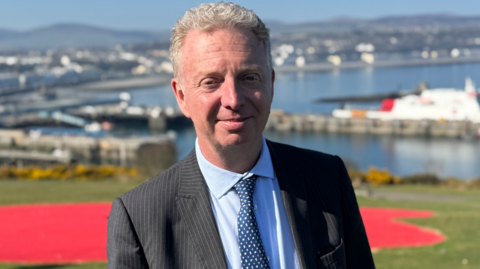 Alex Allinson is wearing a suit, with a black pinstripe jacket, a light blue shirt, and a dark blue spotted tie. He has grey, short, wavy hair and is standing outside on a sunny day, with Douglas Bay behind him.
