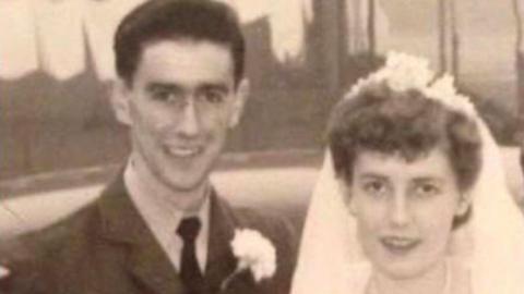 A vintage black-and-white wedding photograph.
In the foreground,Ken and Kate Johnson are standing side by side. Ken is wearing a formal military-style uniform with a belt and decorative buttons, while Kate is dressed in a traditional wedding outfit, including a veil and holding a bouquet of flowers.
