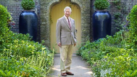 Charles III stands on a garden path leading to a yellow gate in a brick wall. There is well-kept foliage on either side of him and he is wearing a beige blazer, trousers and brown shoes.