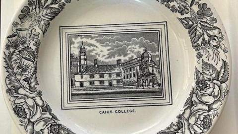 A white plate is decorated with a view of Caius Court in its centre, in black, and the words "Caius College" printed beneath. Around the edges are black drawings of flowers and foliage