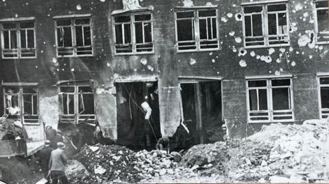 A black and white photograph of a building damaged by bombing. There is debris on the ground and holes in the walls.