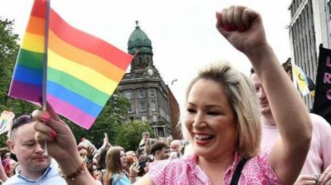 Michelle O'Neill has blonde hair and is wearing a pink t-shirt. She is smiling at the crowd and waving a rainbow Pride flag and pumping her fist while walking among lots of people at a Pride parade in Belfast