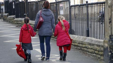 A generic picture showing the back of a woman walking along a street holding hands with two little girls, one with dark hair and the other with blonde hair. The girls are wearing school uniforms under red jackets. The woman is wearing a grey jacket and blue jeans. She has dark hair in a ponytail and is carrying a handbag. All have their backs to the camera. This is a stock picture