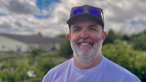 Damien Johnston standing outside with shrubs behind him. He is wearing a baseball cap, with dark glasses propped on its brim and a white T-shirt with Wembley 2 Wigmore written on it. He has a short cropped white beard and is smiling.