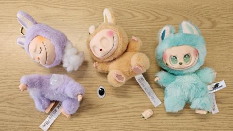 Three dolls that look like monsters sit on a brown surface. A purple doll has it's head disconnected, a cream on has had its eye removed and a turquoise one is missing a foot.