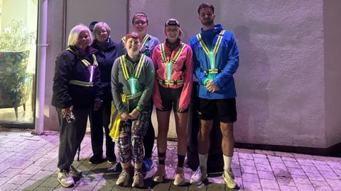 Five women and one man standing outside a sports centre, dressed in running clothes and trainers, and hi-vis vests that are glowing yellow and silver