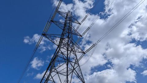 An overhead electricity pylon with cables attached against a blue sky with some clouds