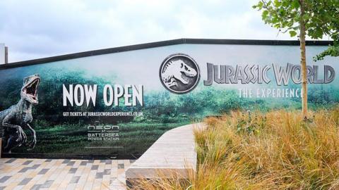 The exterior of the exhibition. There are billboards advertising the Jurassic World experience with pictures of dinosaurs.
