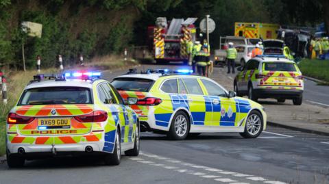 Two police cars with more emergency service vehicles in the background