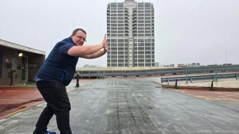 Daniel Adams is using his hands to make it look like he is pushing a building. He is standing in a multi-storey car park. The building is in the middle of the image. Daniel is wearing dark clothing.
