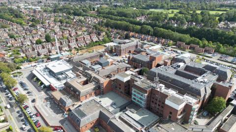 An aerial view of Scunthorpe with a large hospital complex in the foreground, which is made up of multiple connected brick buildings with flat roofs. Roads, parked cars, and service areas surround the site. Beyond the hospital, rows of houses stretch into the distance, bordered by a dense line of trees and green fields.