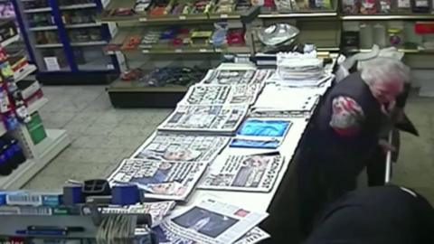A still image of CCTV of a newsagent with papers on the counter and shelves with confectionary for sale. To the right of the counter, a woman in a black gilet with grey hair wields a stick at a man in black who appears to be bent over with his back to her.