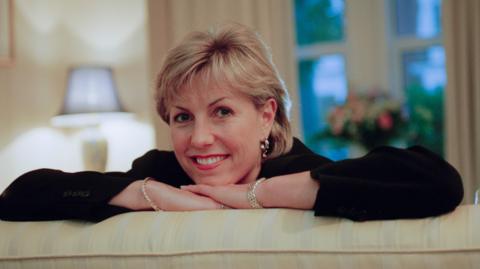 Jill Dando. She has short blonde hair and is wearing a black top and silver jewellery. She is leaning on the back of a sofa, with her chin resting on her hand. She is smiling at the camera. 