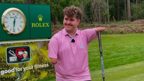 Adam has under-developed arms. He is holding onto a golf club with his arm and wearing a pink golf shirt.