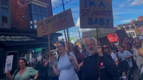 Group of people, from children to older people, walking through town centre hold placards calling for St Mary's  to be saved.
