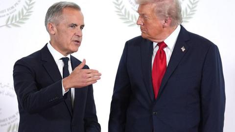 President Donald Trump greets Canada's Prime Minister Mark Carney during a world leaders' summit on ending the Gaza war on October 13, 2025 in Sharm El-Sheikh, Egypt. Prime Minister Mark Carney is on the left, wearing a black tie and a black suit. President Trump is on the right, wearing a dark blue suit and a red tie. PM Carney is gesturing as he speaks to Trump, who appears to be listening. Behind them is a white backdrop. 